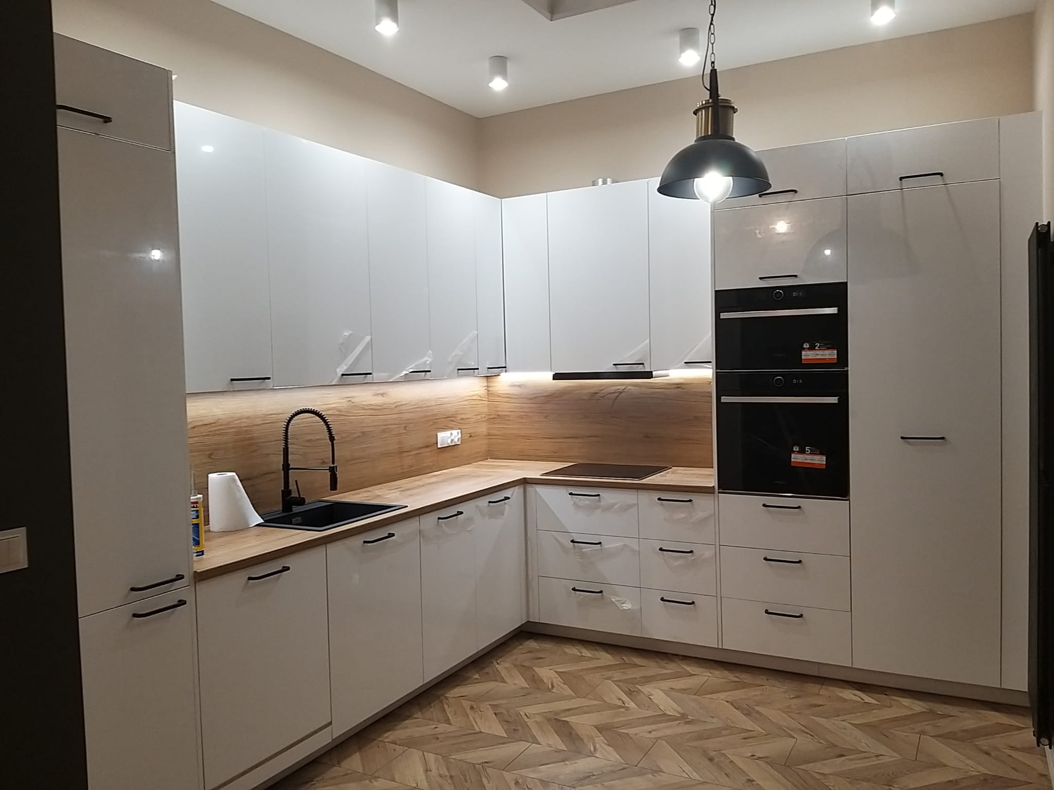 Nowoczesna kuchnia z białymi, lakierowanymi szafkami, drewnianym blatem i podłogą ułożoną w jodełkę, oświetlona wiszącą lampą w stylu industrialnym.