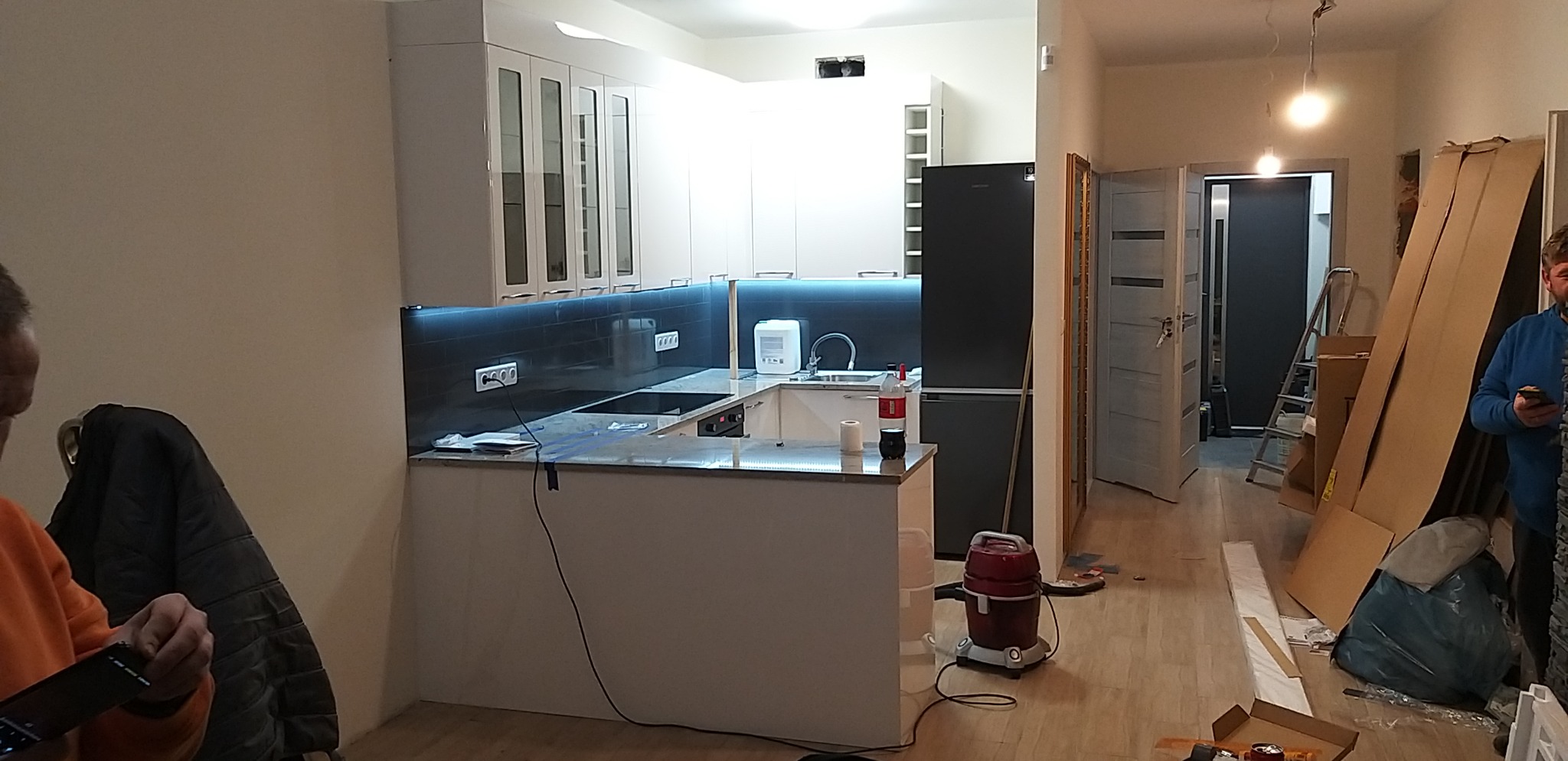 Wnętrze mieszkania w trakcie remontu: widoczna nowa, biała zabudowa kuchenna z podświetleniem LED nad blatem, czarna lodówka, oraz elementy wykończeniowe w korytarzu, w tle widoczna drabina i kartony.