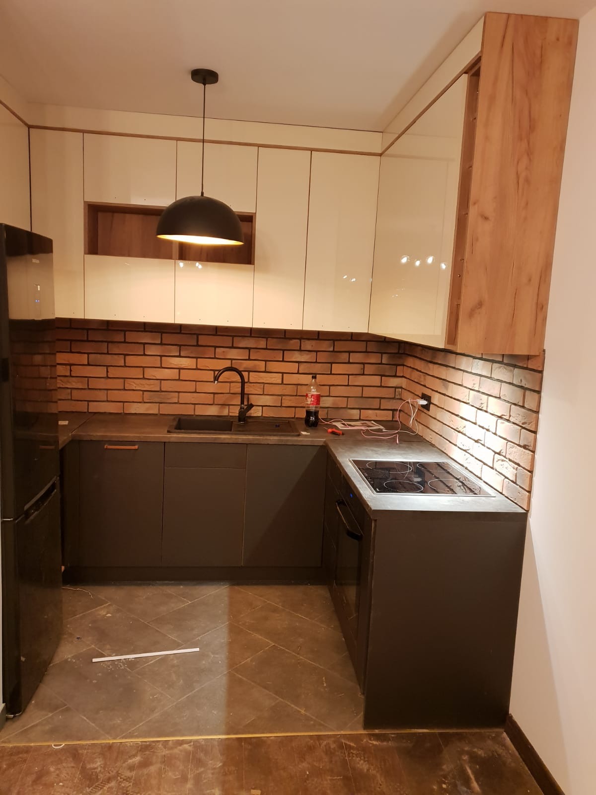 Nowoczesna kuchnia w trakcie wykańczania: szare szafki dolne, białe i drewnopodobne górne, ceglana ściana nad blatem, czarna armatura i lampa wisząca, widoczne przewody elektryczne i butelka napoju...