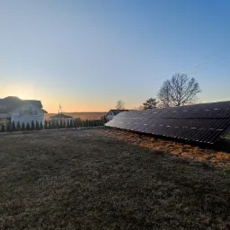 Instalacja paneli słonecznych na trawiastym terenie przed domem jednorodzinnym o wschodzie słońca, widoczne metalowe podpory konstrukcji paneli.
