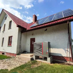 Biały dom jednorodzinny z czerwoną dachówką, panelami fotowoltaicznymi na dachu i pompą ciepła na zewnątrz, z widoczną rzeźbą ptaka na ścianie.