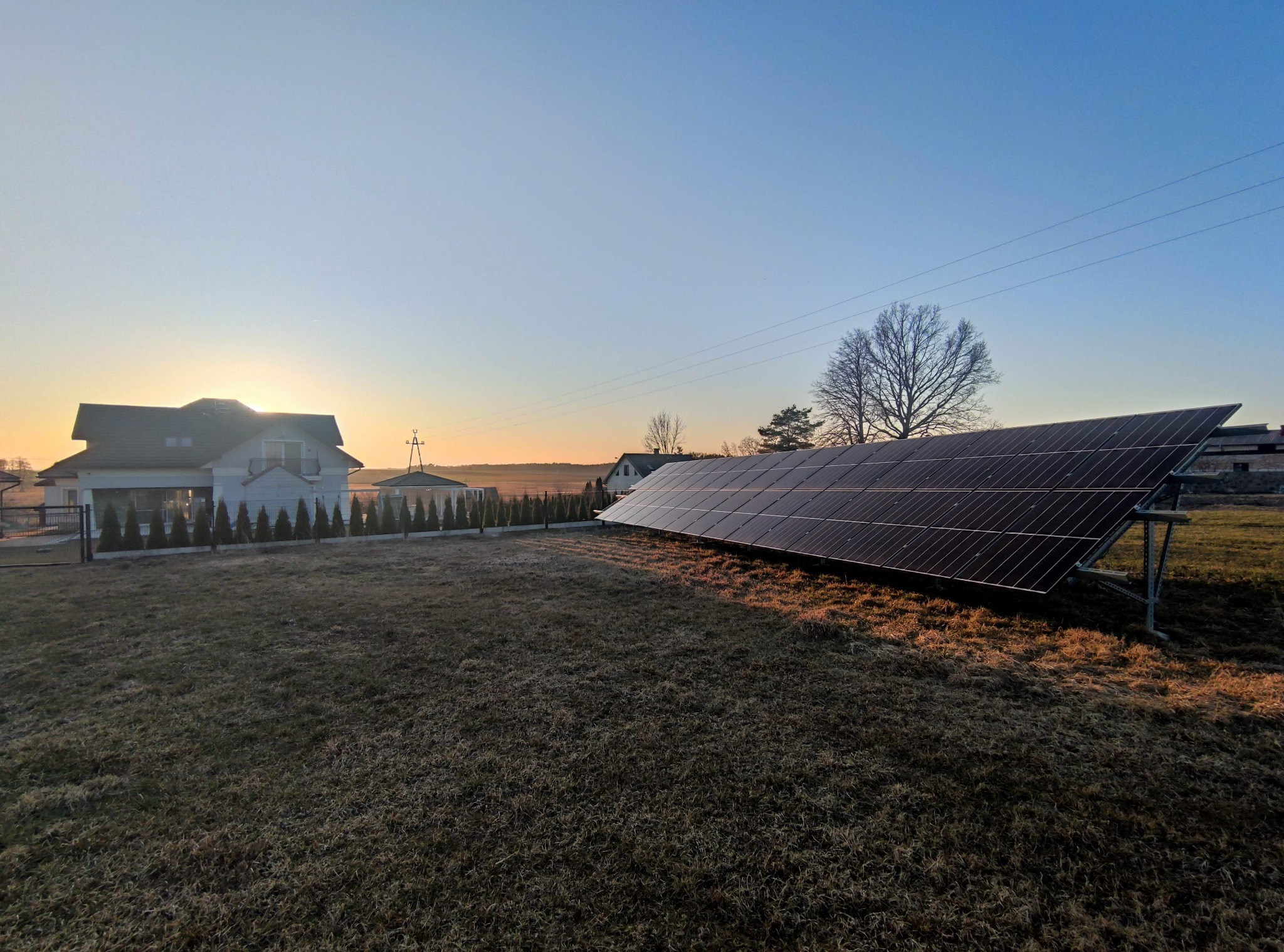 Instalacja paneli słonecznych na trawiastym terenie przed domem jednorodzinnym o wschodzie słońca, widoczne metalowe podpory konstrukcji paneli.