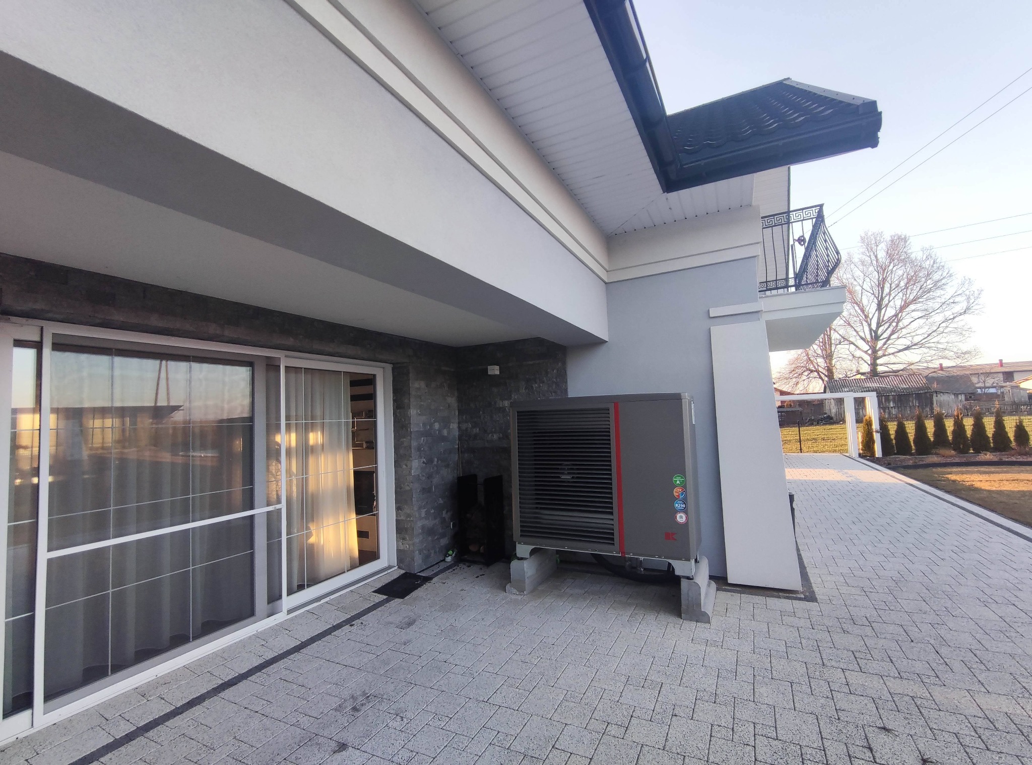 Szara pompa ciepła zainstalowana na betonowych podstawach przy ścianie domu z przesuwnymi drzwiami tarasowymi, widok z zewnątrz na kostce brukowej.