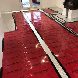 Rozłożone czerwone folie grzewcze marki Dream Heat na srebrnej izolacji podłogowej w jasnym pomieszczeniu z oknem balkonowym i kominkiem.