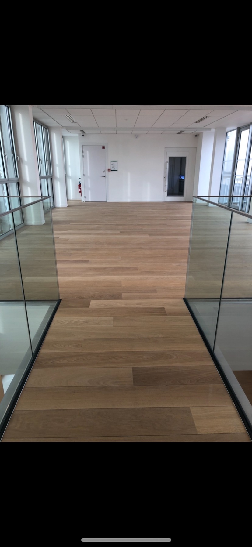 Nowoczesne wnętrze biurowe z drewnianą podłogą, szklanymi balustradami i białymi ścianami, widok z góry na przestrzeń z drzwiami i oknami.