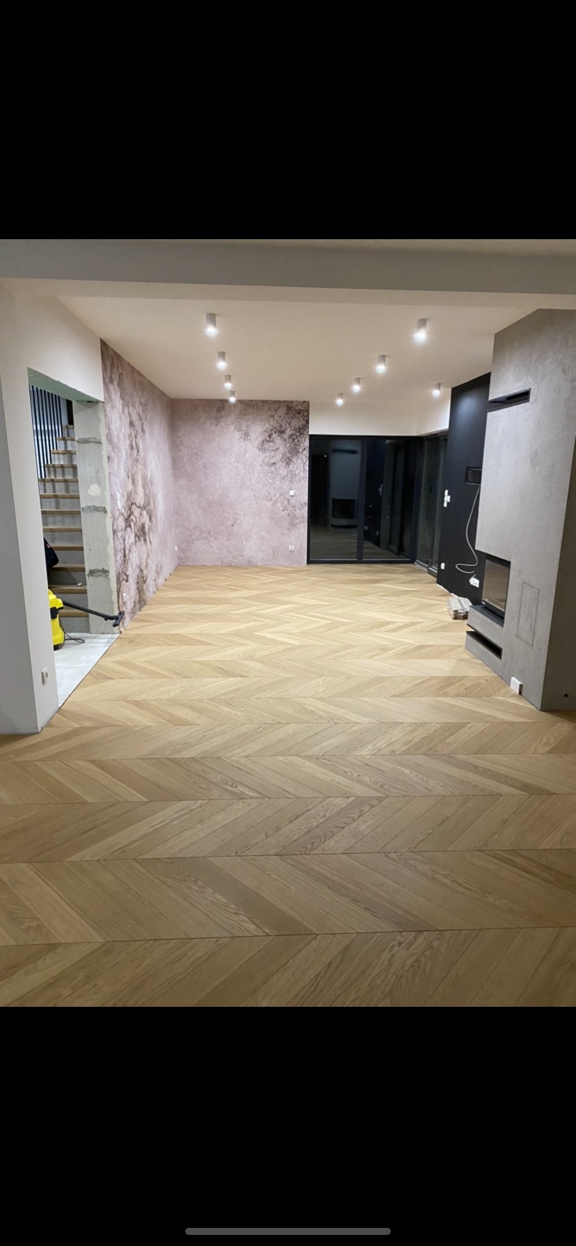 Salon z nową podłogą parkietową w jodełkę, ściana z tapetą imitującą marmur, nowoczesny kominek i surowe schody w trakcie wykończenia.