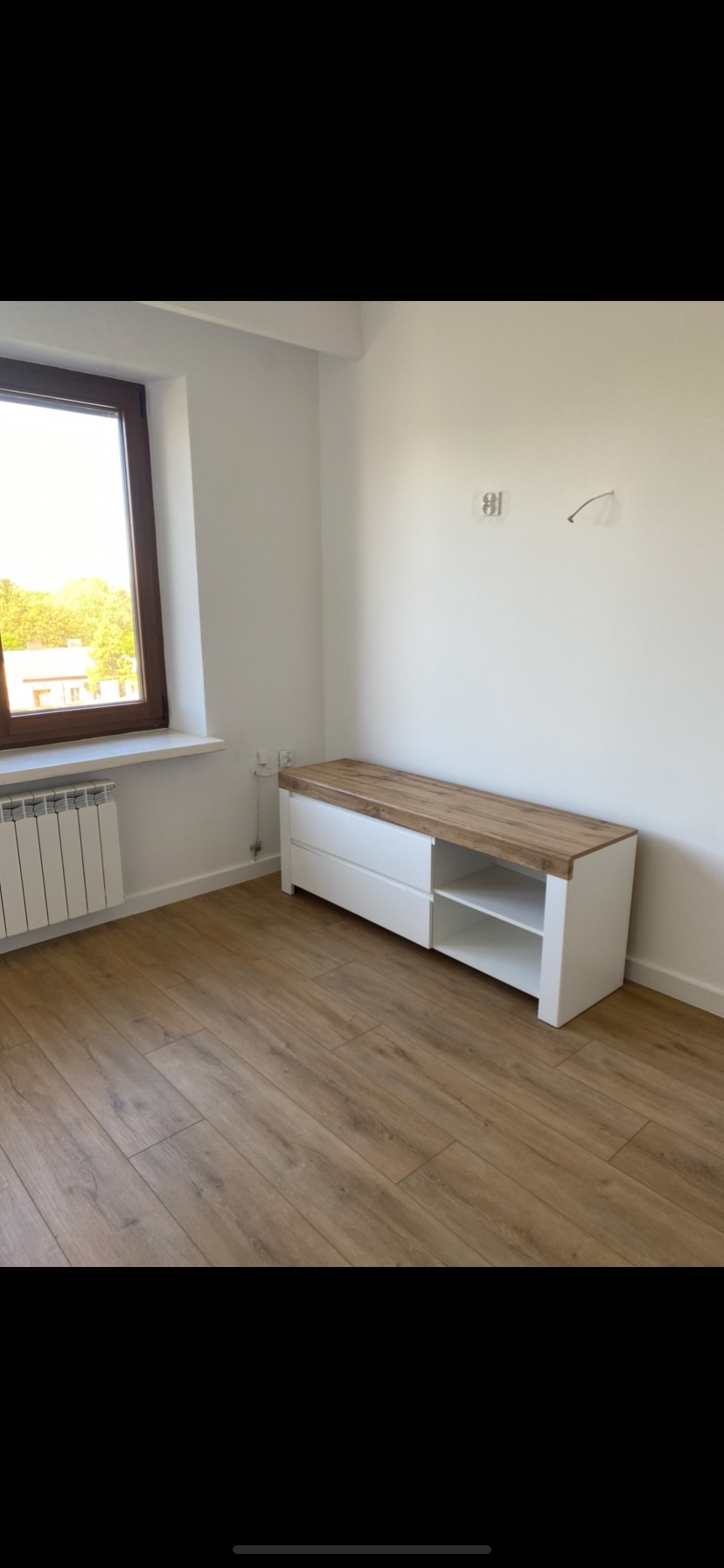 Pusta, biała ściana w pokoju z drewnianą podłogą, grzejnikiem pod oknem i białą szafką z drewnianym blatem.