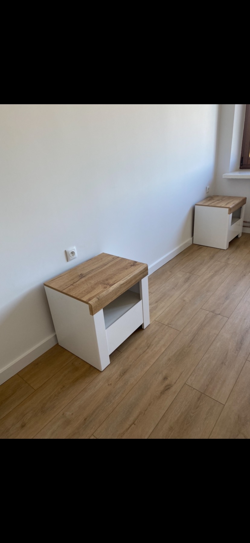 Dwie białe szafki nocne z drewnianymi blatami na podłodze z paneli w jasnym pokoju, widoczny kontakt na ścianie.