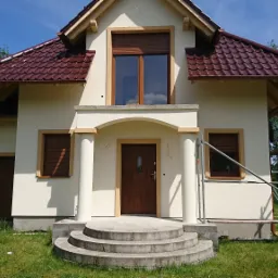 Jasny dom jednorodzinny z brązową dachówką i roletami zewnętrznymi, widoczne rusztowanie i drabina malarska