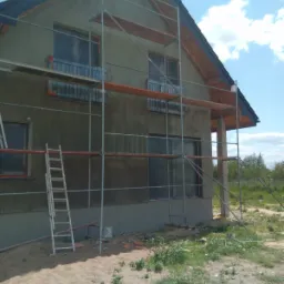 Dom jednorodzinny w trakcie prac elewacyjnych, widoczne rusztowania, zabezpieczone okna folią i świeżo nałożony tynk w szarym odcieniu.