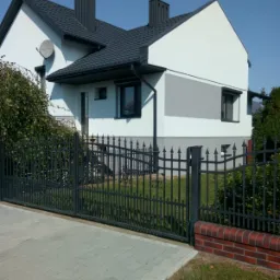 Dwukondygnacyjny dom z białą elewacją z szarym pasem, ciemną dachówką, czarną metalową bramą i brukowanym podjazdem w słoneczny dzień.