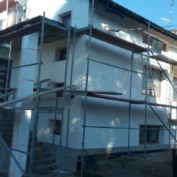 Dwupiętrowy dom w trakcie ocieplania, pokryty białymi płytami styropianowymi, otoczony metalowymi rusztowaniami, z widocznym fragmentem dachu i ogrodu w tle.