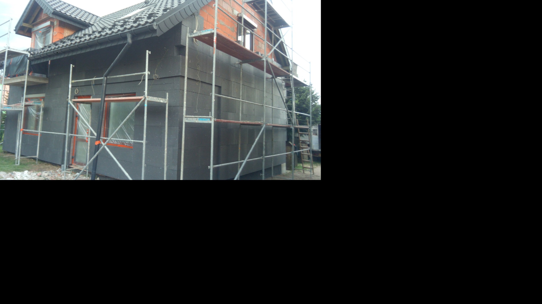 Dom jednorodzinny w trakcie ocieplania, ściany pokryte czarnym styropianem, rusztowania wokół budynku, okna zabezpieczone folią.