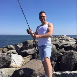 Mężczyzna w okularach, białym podkoszulku i krótkich jeansowych spodenkach, stojący na kamiennym nabrzeżu z wędką w rękach, na tle morza i błękitnego nieba.