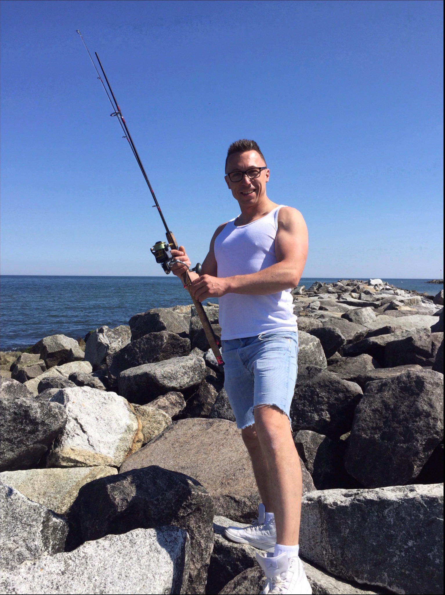 Mężczyzna w okularach, białym podkoszulku i krótkich jeansowych spodenkach, stojący na kamiennym nabrzeżu z wędką w rękach, na tle morza i błękitnego nieba.