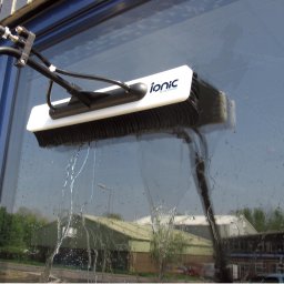 KOSTRADE Jarosław Kostka - Szczotka teleskopowa z logiem Ionic Systems czyści szklaną elewację budynku, w tle widoczne budynki przemysłowe.