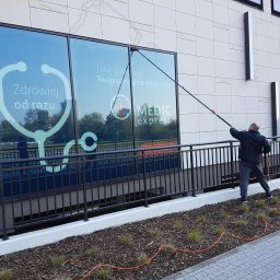 KOSTRADE Jarosław Kostka - Pracownik w trakcie czyszczenia okien budynku Medic Express za pomocą teleskopowej lancy z dyszą, widoczny pomarańczowy wąż doprowadzający wodę.
