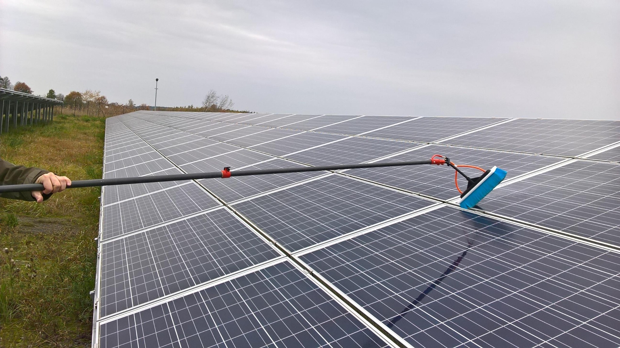 Czyszczenie paneli słonecznych za pomocą szczotki na długim wysięgniku; widoczny operator trzymający wysięgnik i rząd paneli w tle.