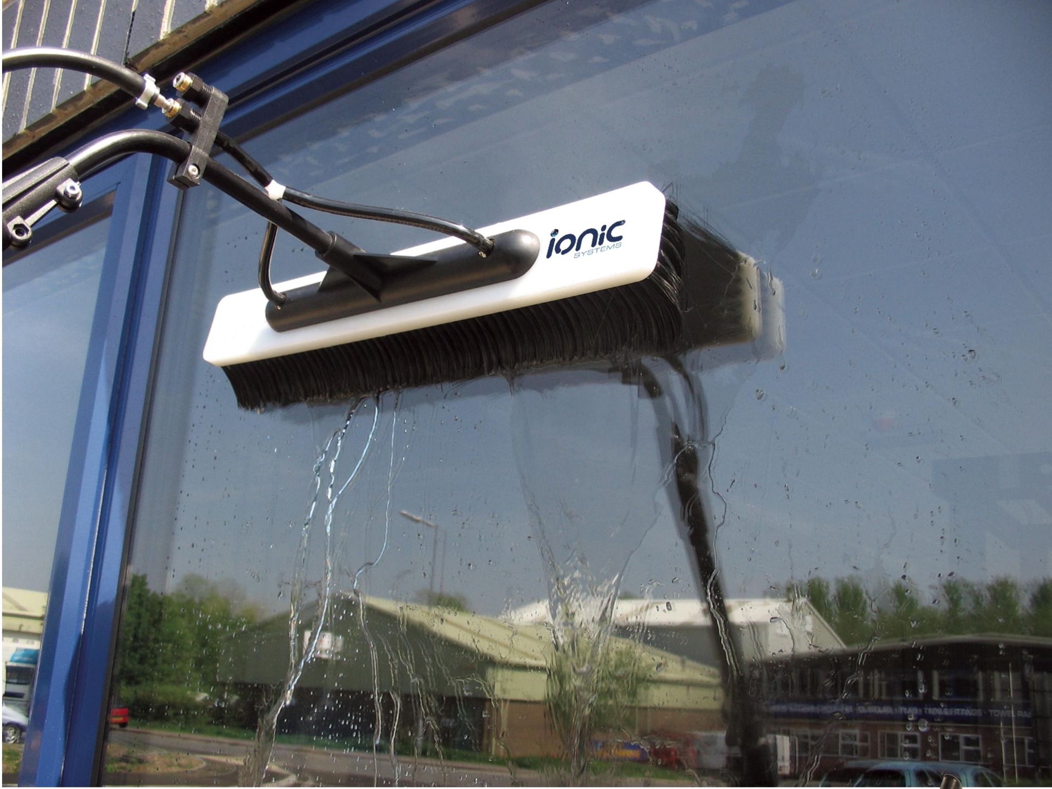 Szczotka teleskopowa z logiem Ionic Systems czyści szklaną elewację budynku, w tle widoczne budynki przemysłowe.