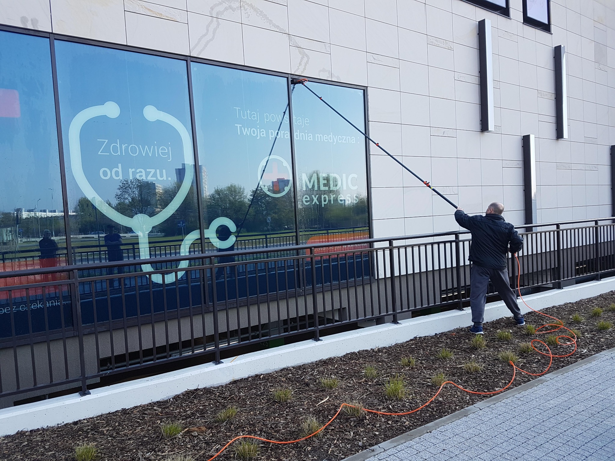 Pracownik w trakcie czyszczenia okien budynku Medic Express za pomocą teleskopowej lancy z dyszą, widoczny pomarańczowy wąż doprowadzający wodę.