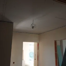 Wnętrze w trakcie remontu: sufit z płyt gipsowo-kartonowych z widocznymi śrubami, ściany przygotowane do dalszych prac, widoczne kable oświetleniowe i otwór w suficie.