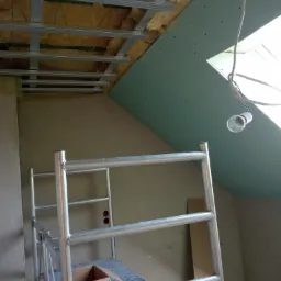 Szkielet metalowej konstrukcji podwieszanego sufitu z widoczną izolacją z wełny mineralnej na poddaszu, fragment rusztowania i lampa wisząca.