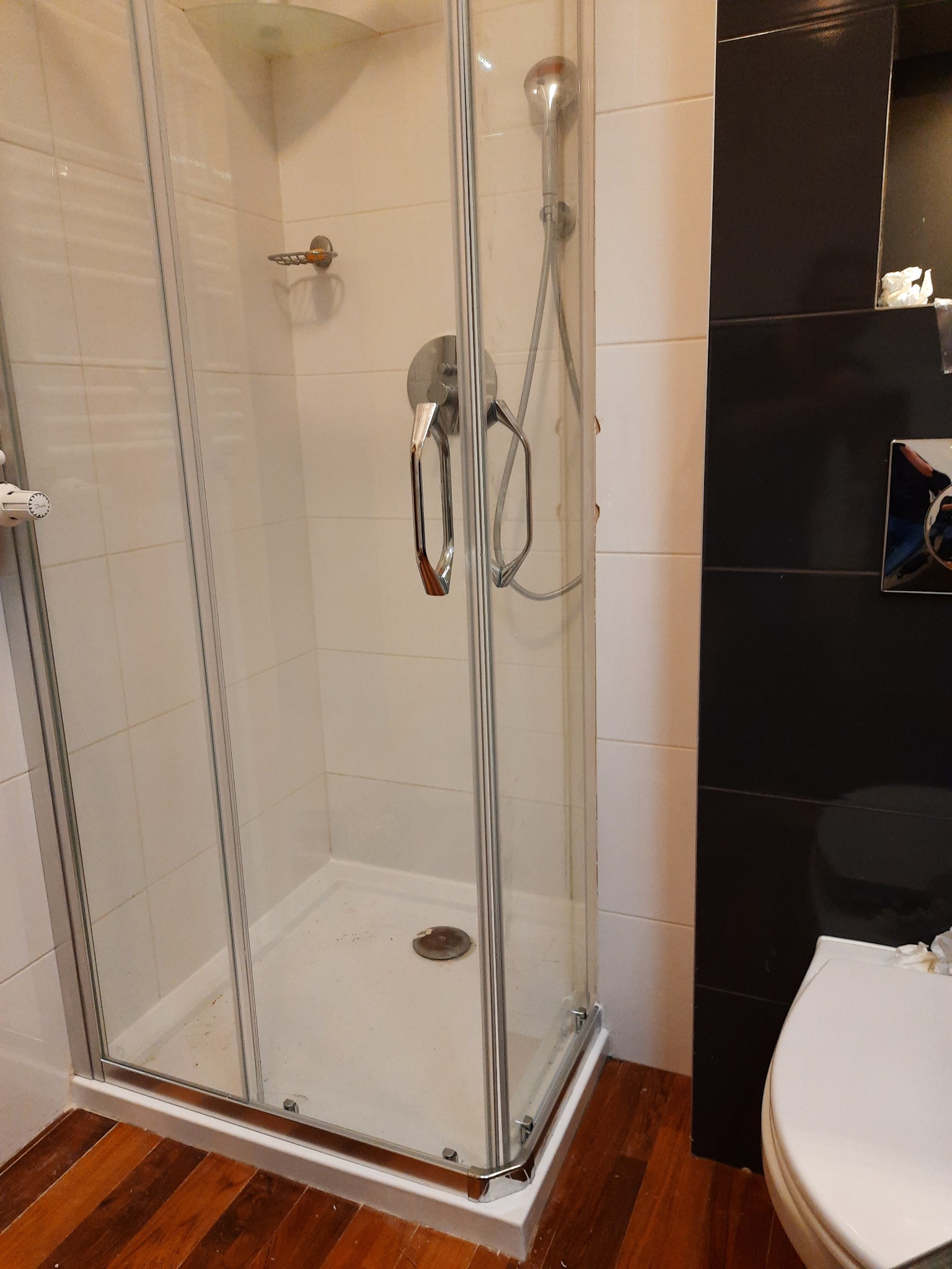 Kabiną prysznicowa z chromowaną armaturą, zamontowana w łazience z drewnianą podłogą i czarnymi płytkami, widoczne ślady użytkowania.