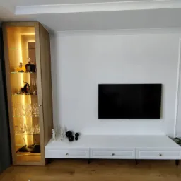 Nowoczesny salon z podświetlaną witryną z drewnianym wykończeniem, białą szafką RTV z trzema szufladami i czarnym telewizorem na białej ścianie. Na półkach witryny szklanki i butelki z alkoholem.