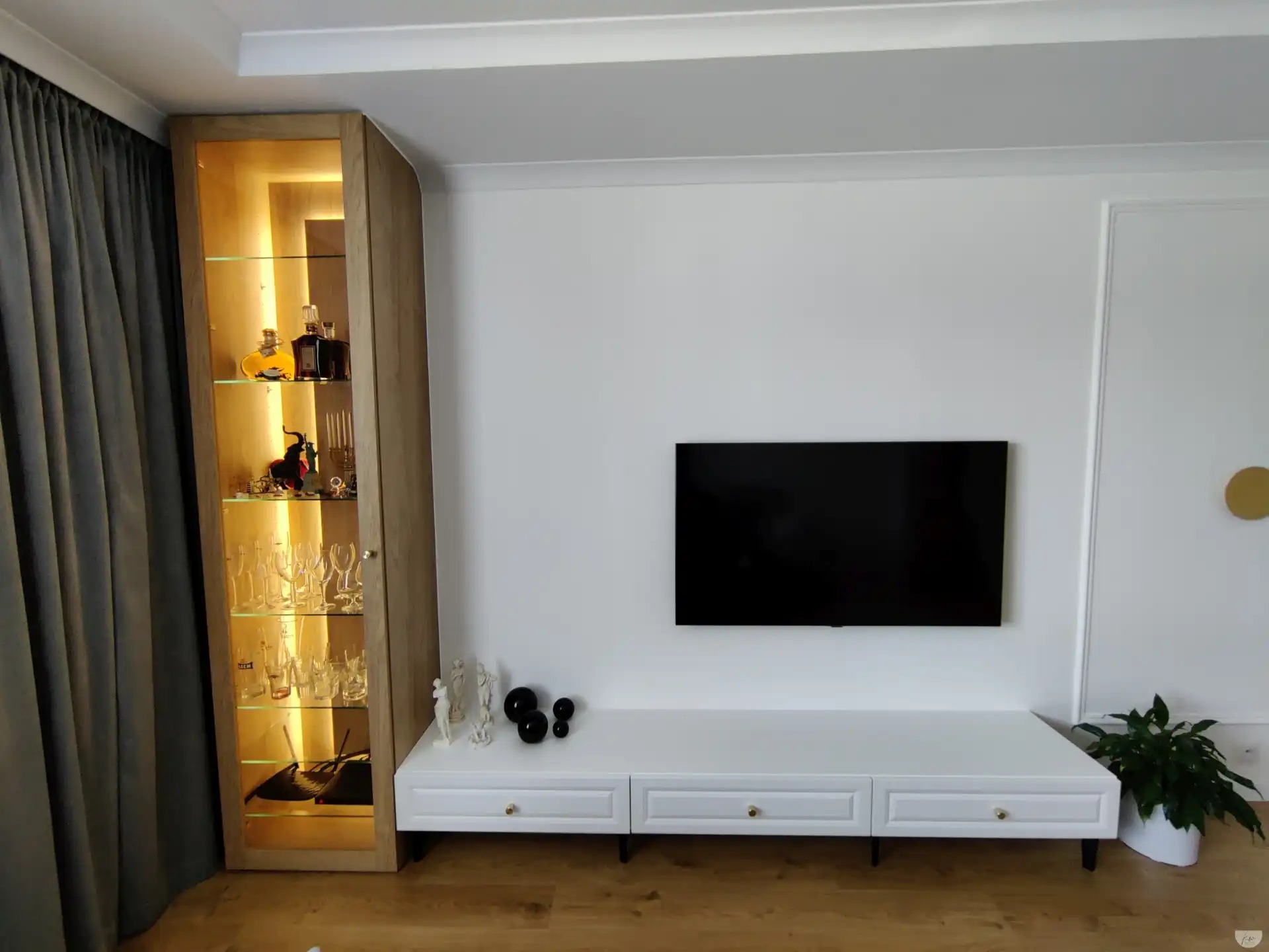Nowoczesny salon z podświetlaną witryną z drewnianym wykończeniem, białą szafką RTV z trzema szufladami i czarnym telewizorem na białej ścianie. Na półkach witryny szklanki i butelki z alkoholem.
