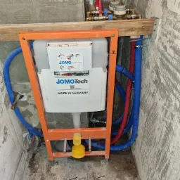 Stelaż podtynkowy WC JomoTech w pomarańczowej ramie, widoczne przyłącza wody, nieotynkowane ściany w trakcie prac remontowych.