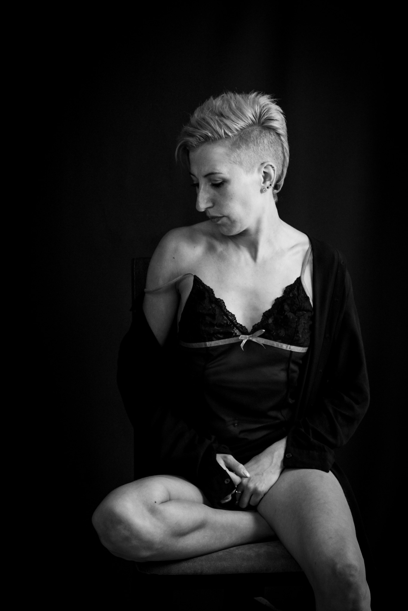 Portret kobiety w bieliźnie, siedzącej na krześle na czarnym tle, z krótkimi włosami i kolczykami w uszach, w czarno-białej tonacji.