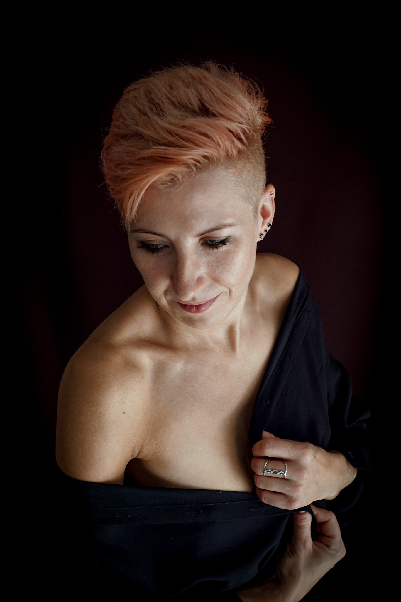 Portret kobiety z krótkimi, brzoskwiniowymi włosami, spoglądającej w dół, ubranej w czarną koszulę z odsłoniętym ramieniem, z delikatnym uśmiechem i pierścionkiem na palcu.