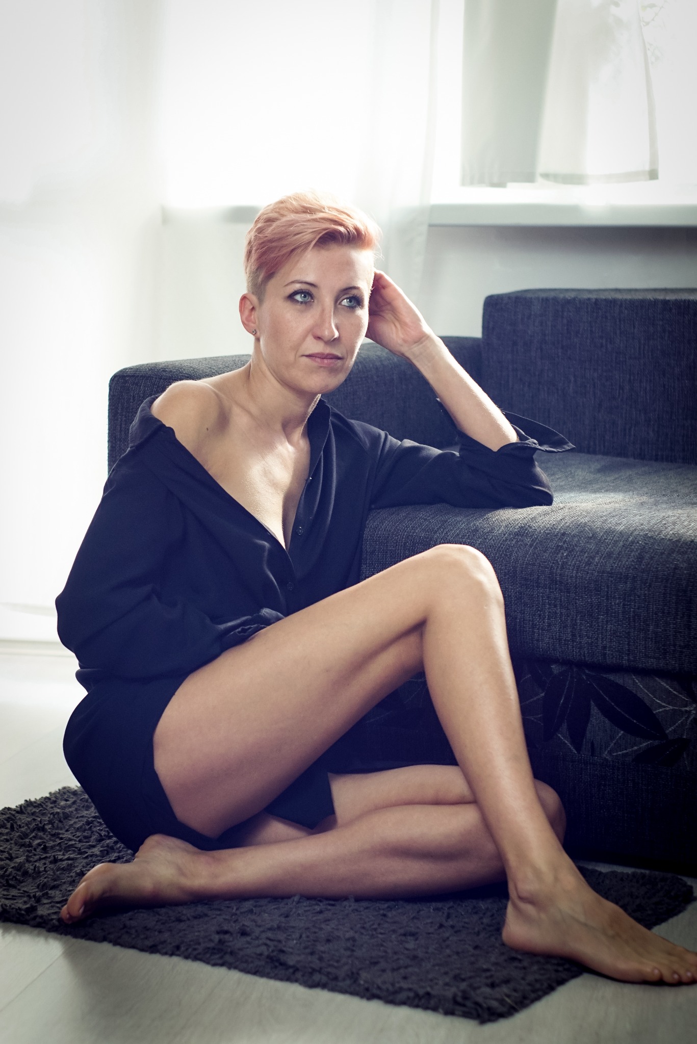 Kobieta z krótkimi, różowymi włosami siedzi na dywanie, ubrana w rozpiętą, czarną koszulę, opierając głowę o rękę i patrząc w dal.