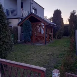 Szara, dwupiętrowa bryła domu z balkonem i drewnianą altaną w ogrodzie, widziana zza zardzewiałego, metalowego ogrodzenia.