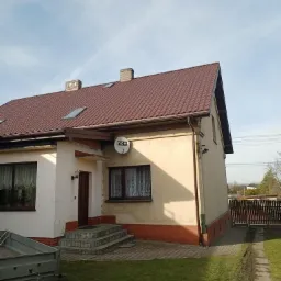 Dom jednorodzinny z brązowym dachem z dachówki, widoczne dwa kominy, okna dachowe i antena satelitarna na elewacji.