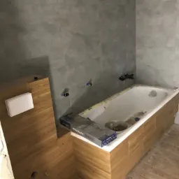 Wanna obudowana panelami imitującymi drewno, szare płytki na ścianie, widoczne fragmenty instalacji wodnej, paczka płytek Moza Black na obudowie wanny, podłoga częściowo wyłożona panelami.