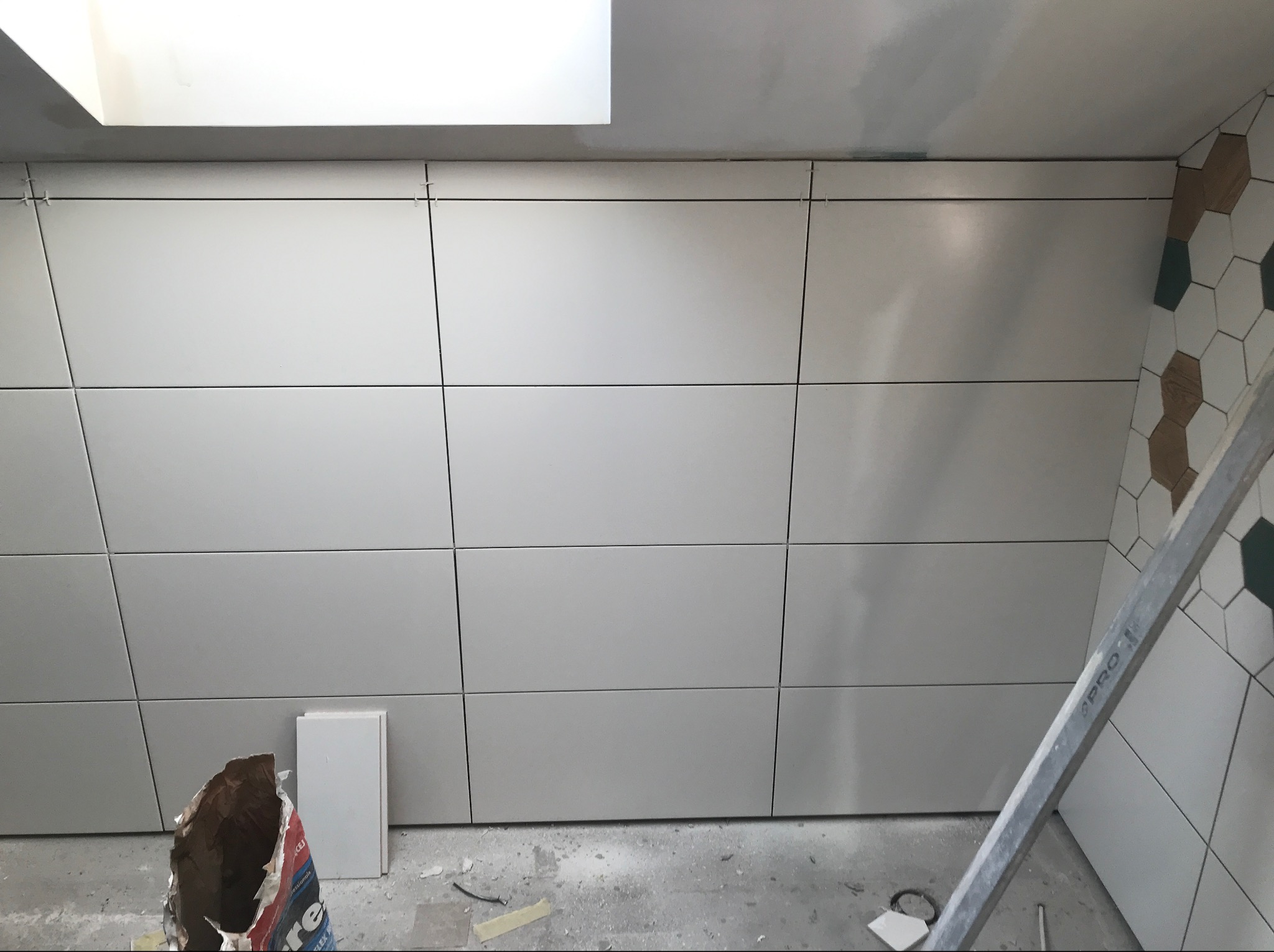 Ściana wyłożona dużymi, matowymi, szarymi płytkami w trakcie montażu, widoczne krzyżyki dystansowe, resztki zaprawy i drabina oparta o ścianę z fragmentem dekoracyjnej mozaiki heksagonalnej w rogu.
