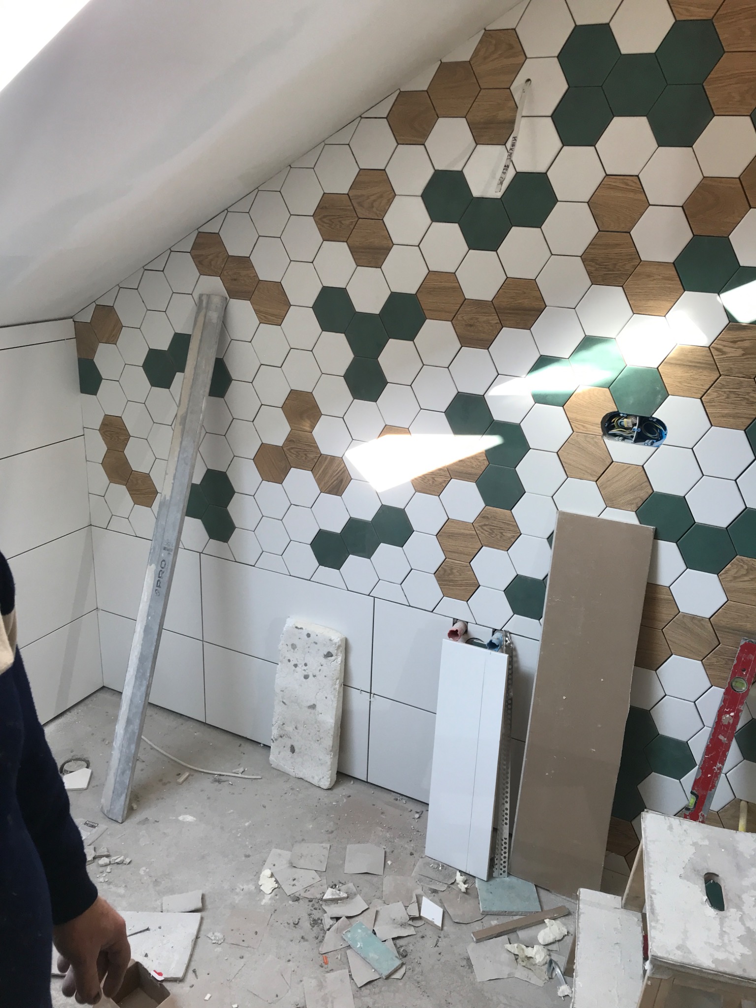 Ściana w łazience wyłożona heksagonalnymi płytkami w kolorach białym, zielonym i imitującym drewno, widoczny fragment instalacji elektrycznej, poziomica i resztki materiałów budowlanych na podłodze.