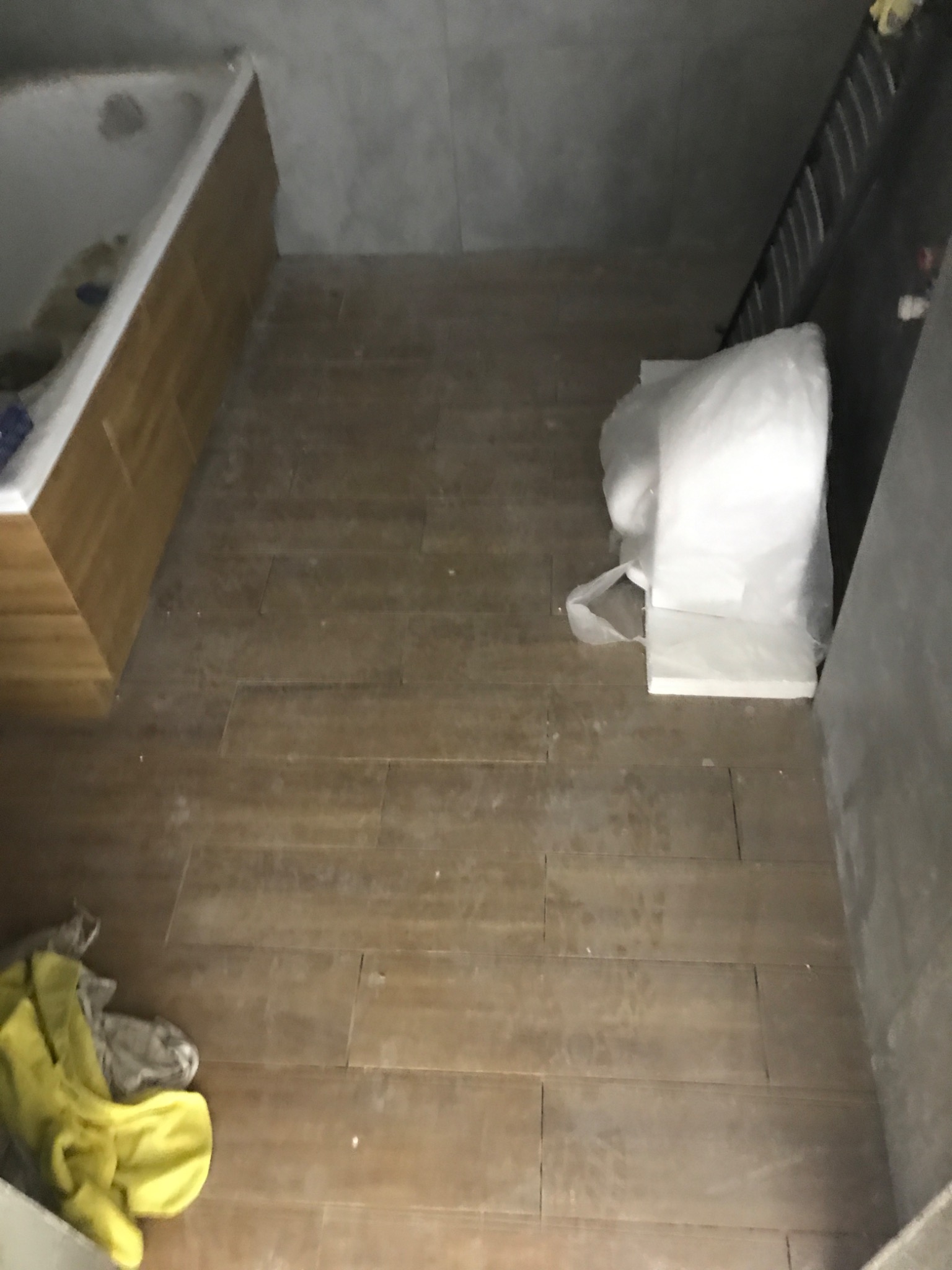 Brązowe płytki podłogowe imitujące drewno w trakcie układania, widoczna wanna z obudową, resztki zaprawy i narzędzia glazurnicze w rogu pomieszczenia.