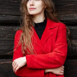 Portret kobiety w czerwonym płaszczu na tle ciemnej, drewnianej ściany, zamyślona poza i delikatny uśmiech.