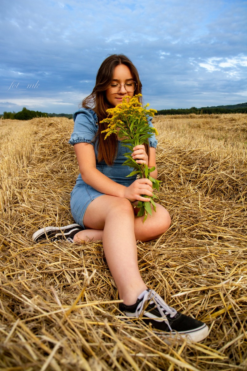 Młoda kobieta w dżinsowej sukience siedzi na polu ze słomą, trzymając bukiet żółtych kwiatów, pochylona twarzą w kierunku kwiatów, ubrana w okulary i czarne trampki.