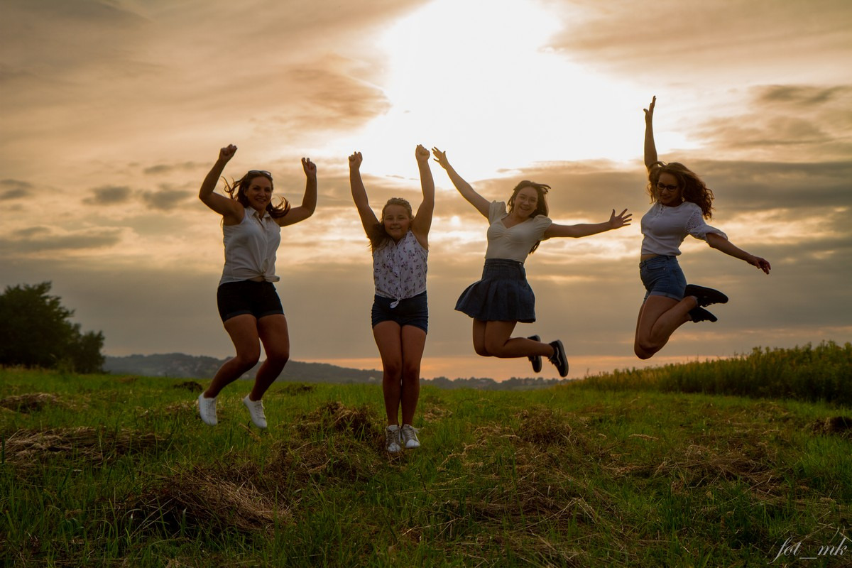 Cztery roześmiane dziewczyny skaczą na łące w promieniach zachodzącego słońca, sylwetki na tle pomarańczowego nieba.