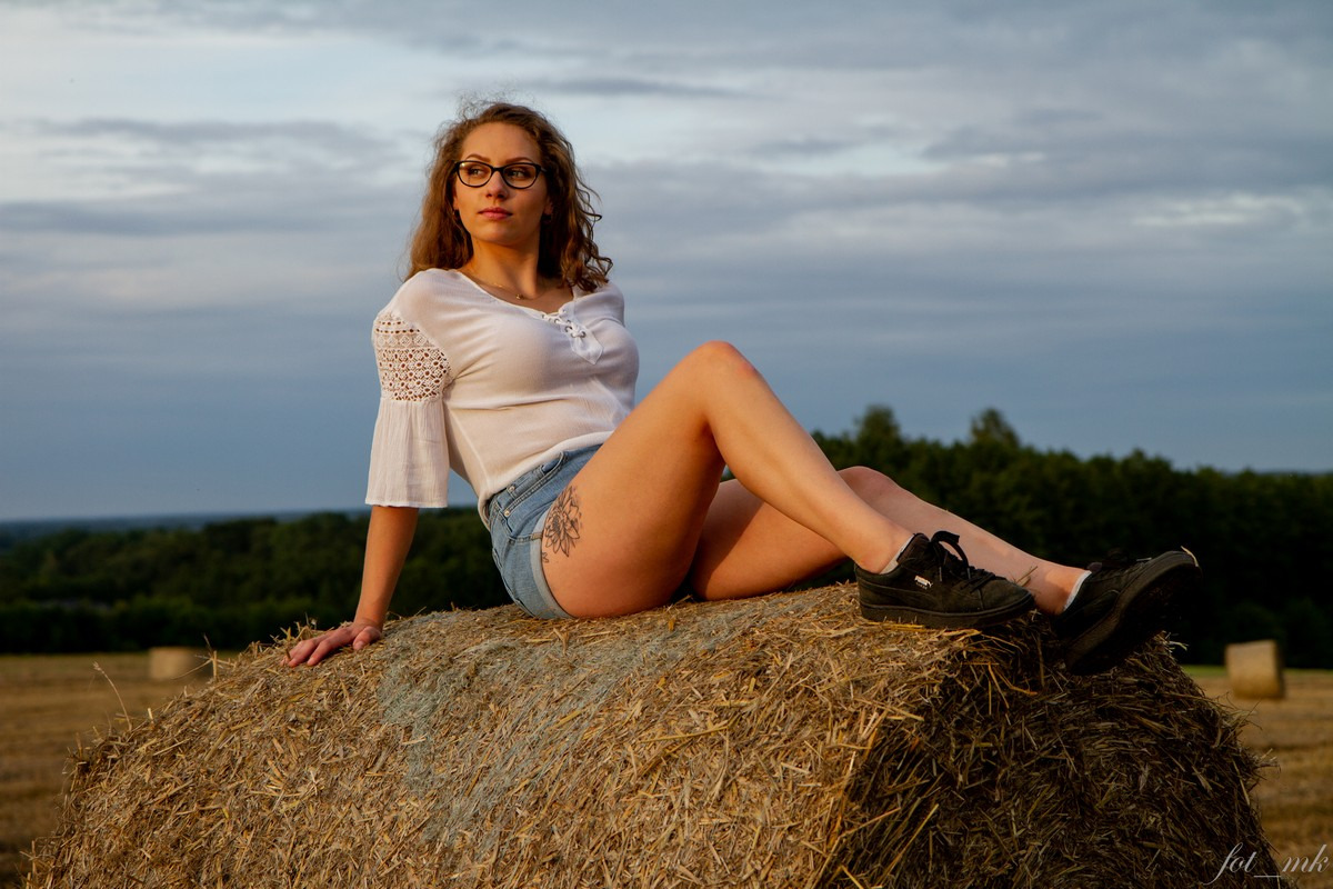 Kobieta w okularach siedzi na belocie siana na tle pola i zachodzącego słońca.