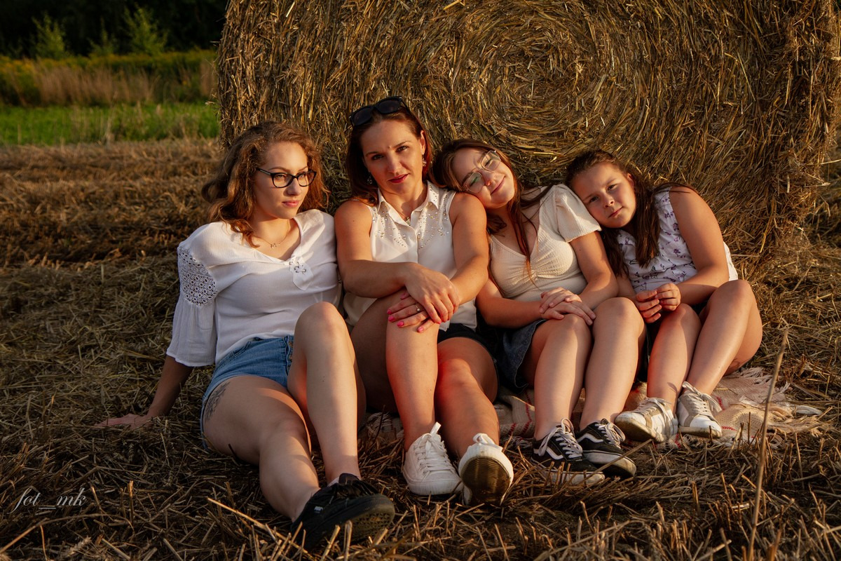 Cztery kobiety w różnym wieku siedzą oparte o okrągły balot słomy na polu w promieniach zachodzącego słońca, ubrane w letnie ubrania, z widocznym podpisem fotografa w lewym dolnym rogu.