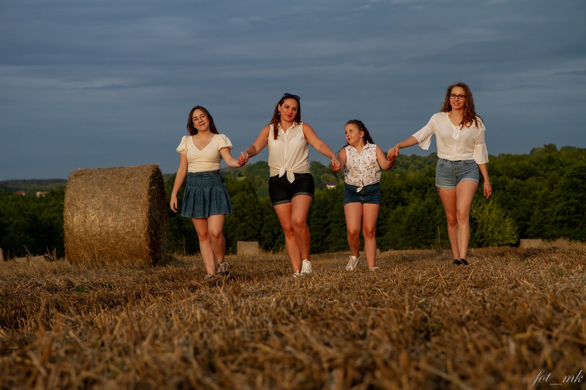 Cztery kobiety trzymają się za ręce, spacerując po polu ze słomą w letni wieczór, w tle widoczne drzewa i zachmurzone niebo, obok leży bela słomy.