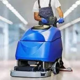 Pracownik w kombinezonie obsługuje niebieską maszynę do czyszczenia podłóg w jasnym korytarzu biurowym, widoczne odblaski na podłodze.