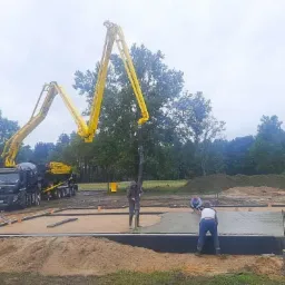 Wylewanie fundamentów pod budynek za pomocą pompy do betonu, widoczni pracownicy budowlani i ciężarówka z betonomieszarką na tle zieleni.
