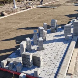 Układanie szarej kostki brukowej na podsypce piaskowej, widoczny pracownik w odblaskowej kamizelce w tle, część kostki ułożona, część w stosach do ułożenia.