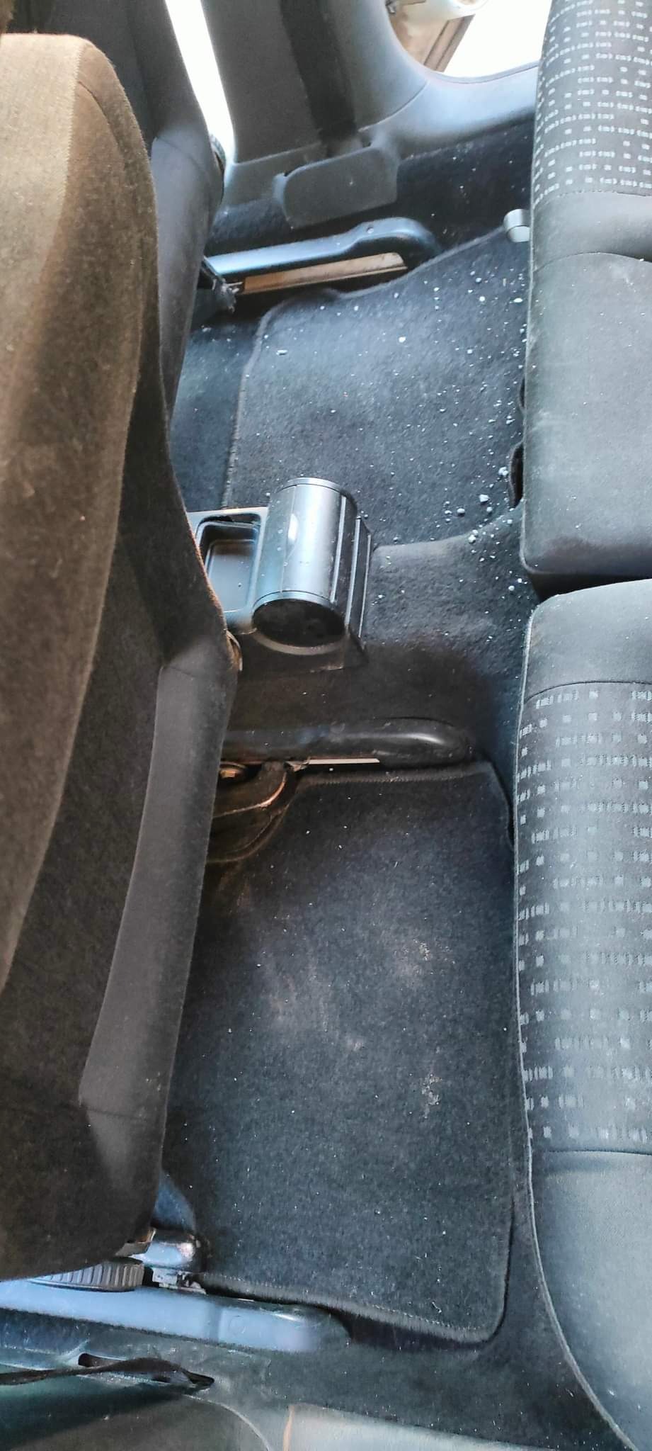 Wnętrze samochodu przed czyszczeniem: brudna, czarna wykładzina podłogowa z widocznymi okruchami i plamami, uchwyt na kubek, fragment siedzenia z wzorem w kratkę.