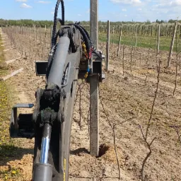 Wbijanie betonowego słupka w ziemię przy użyciu specjalistycznej maszyny w sadzie. W tle widoczne rzędy drzewek i błękitne niebo z chmurami.